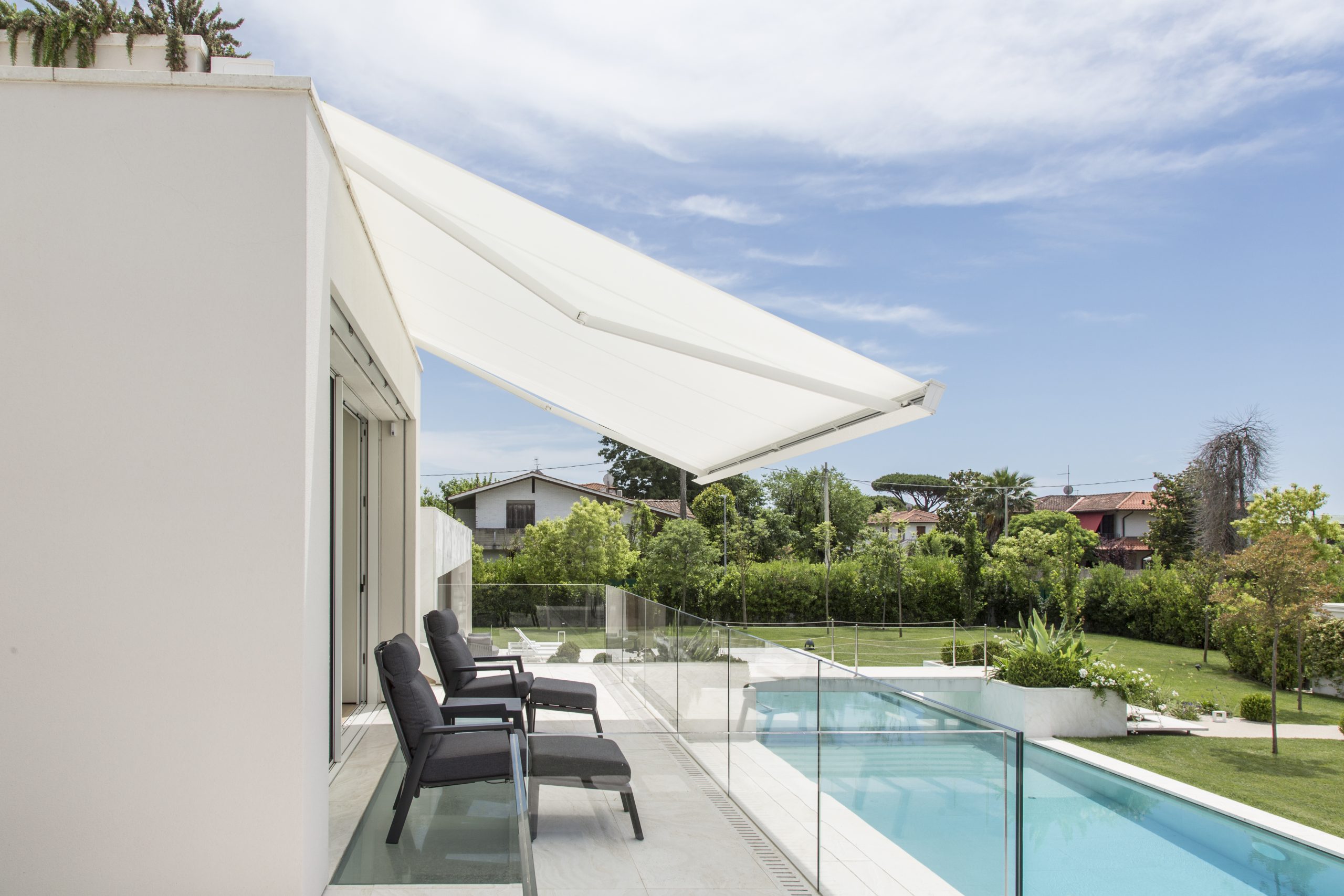 Tenda da sole moderna a bracci estensibili, montata su una terrazza con vista su piscina e giardino, con sedie nere su pavimento bianco.